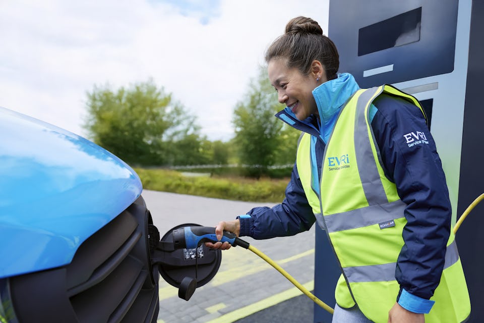 A female van driver dressed in an Evri branded hi-vis vest is charging a blue Evri electric van.